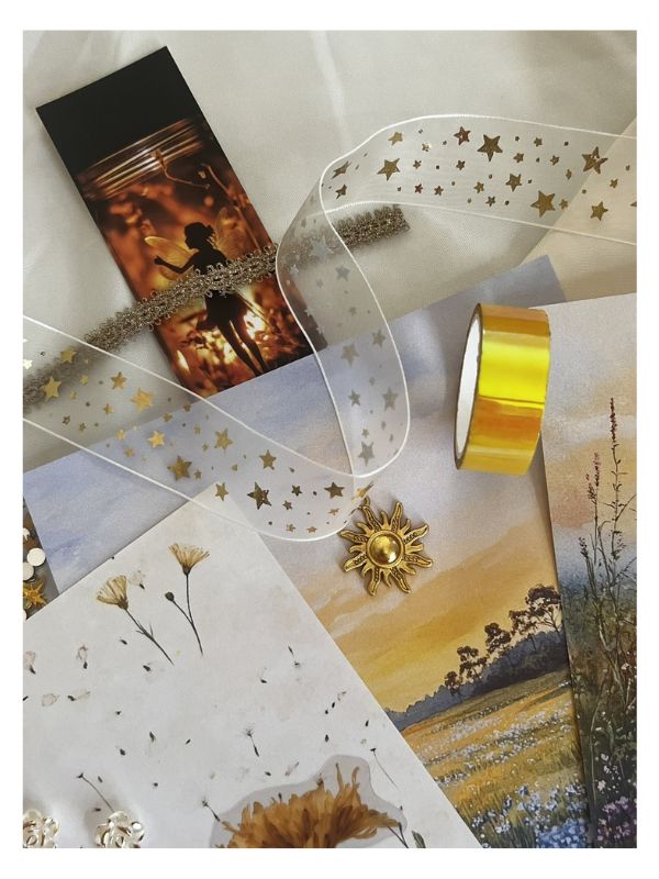 A styled flat-lay of decorative craft supplies on a light fabric background, including a roll of metallic gold tape, a clear ribbon with gold star pattern, a small sun-shaped embellishment, dried flower pieces, and printed cards or paper with scenic artwork.