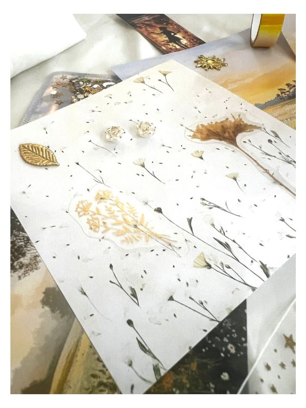 A decorative white card or paper featuring pressed wildflower elements, small dried botanical pieces, and scattered black seed-like specks, photographed on top of other craft or stationery items.
